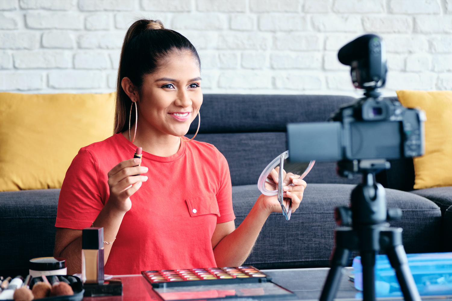  A young woman is sitting in front of a camera and talking while holding a makeup product in her hand.